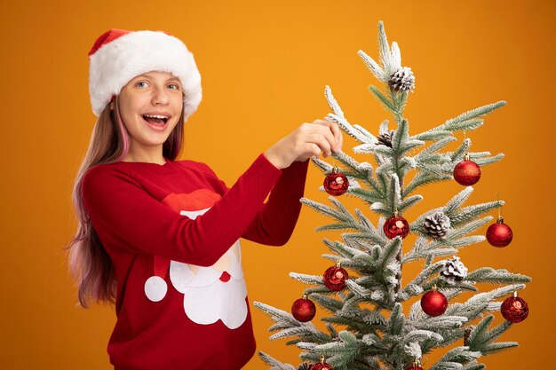 Niña feliz en suéter de navidad y gorro de Papá Noel colgando bolas en un árbol de navidad sonriendo alegremente sobre fondo naranja