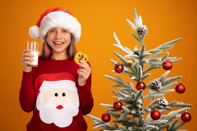 Niña feliz en suéter de Navidad y gorro de Papá Noel con vaso de leche y galletas mirando a la cámara sonriendo alegremente de pie junto a un árbol de Navidad sobre fondo naranja