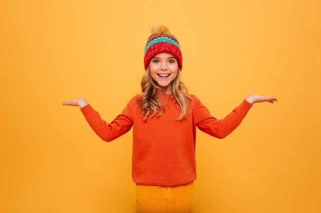 Niña feliz en suéter y sombrero se encoge de hombros mientras mira a la cámara sobre naranja