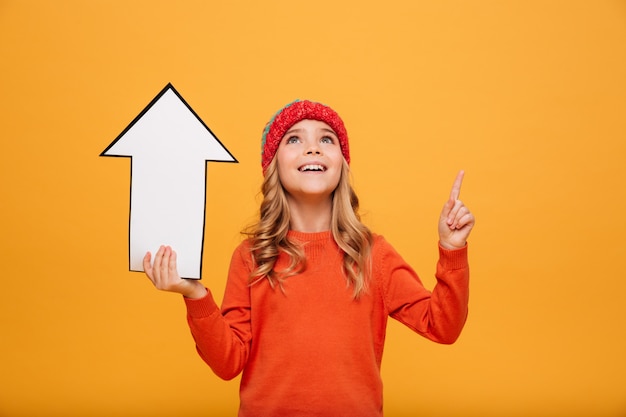 Niña feliz en suéter y sombrero con flecha de papel mientras apunta y mira hacia arriba sobre naranja