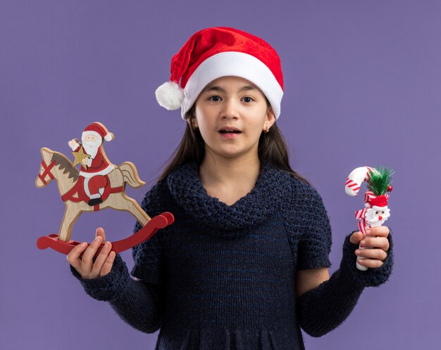 Niña feliz en tejido vestido vistiendo gorro de Papá Noel sosteniendo juguetes de navidad mirando a la cámara sonriendo alegremente de pie sobre fondo púrpura