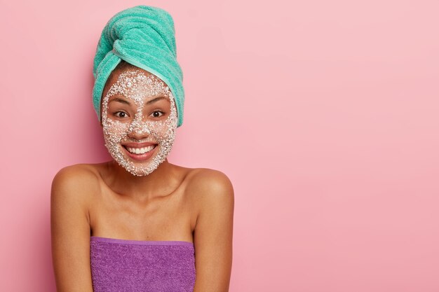 La niña feliz tiene una expresión alegre, se para en el baño después de bañarse, usa un exfoliante alrededor de la cara, envuelta en toallas suaves