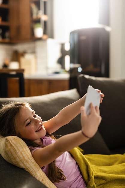 Niña feliz usando teléfono móvil y tomando selfie en el sofá de la sala de estar