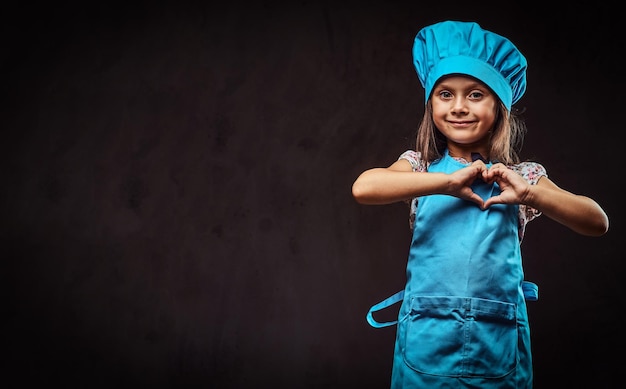 Niña feliz vestida con uniforme de cocinero azul muestra gesto de amor. Aislado sobre fondo oscuro con textura.