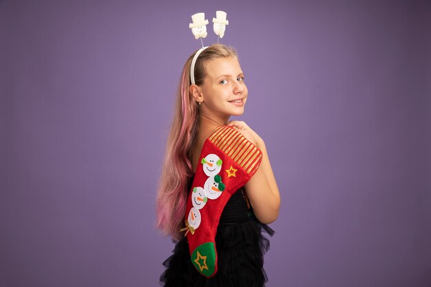 Niña feliz en vestido de fiesta brillante y diadema divertida sosteniendo calcetín de Navidad mirando a la cámara sonriendo alegremente de pie sobre fondo púrpura