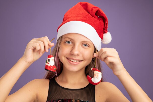 Niña feliz en vestido de fiesta brillante y gorro de Papá Noel con juguetes de Navidad mirando a la cámara sonriendo ampliamente de pie sobre fondo púrpura