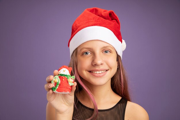 Niña feliz en vestido de fiesta de brillo y gorro de Papá Noel con juguete de Navidad mirando a la cámara sonriendo alegremente de pie sobre fondo púrpura