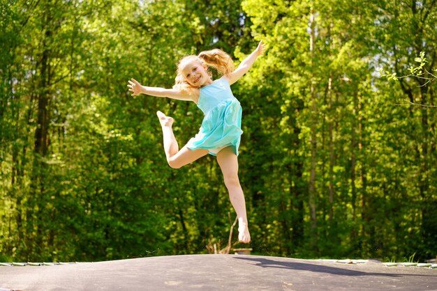 Una niña feliz con un vestido salta sobre un trampolín en un parque en un día soleado de verano