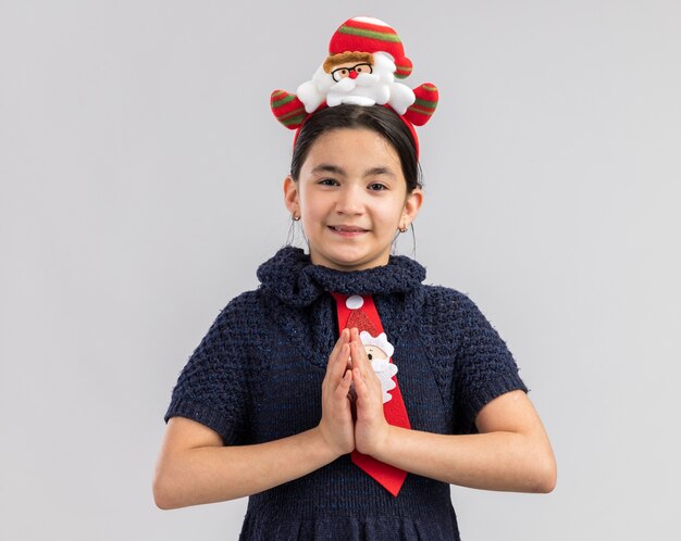 Niña feliz en vestido tejido con corbata roja con divertido borde navideño en la cabeza cogidos de la mano juntos como gesto de namaste