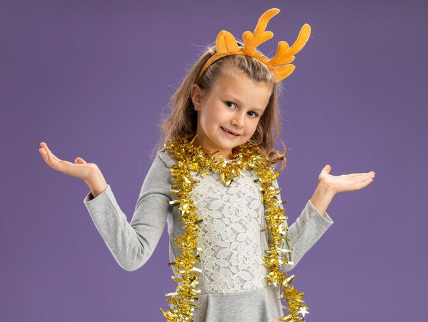 Niña feliz vistiendo un aro de pelo de Navidad con guirnalda en el cuello extendiendo las manos aisladas sobre fondo azul.