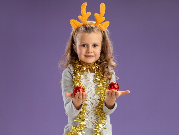 Niña feliz vistiendo un aro de pelo de navidad con guirnalda en el cuello sosteniendo bolas de navidad aislado sobre fondo azul.