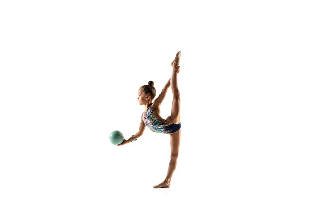 Niña flexible aislada en la pared blanca. Pequeña modelo femenina como artista de gimnasia rítmica en leotardo brillante. Gracia en movimiento, acción y deporte. Haciendo ejercicios con la pelota.