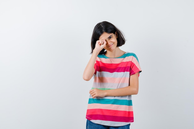 Niña frotándose el ojo en camiseta y mirando feliz. vista frontal.