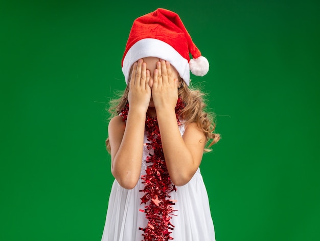 Niña con gorro de navidad con guirnalda en el cuello con la cara cubierta con las manos aisladas en la pared verde