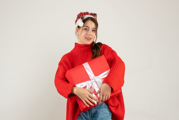 Niña con gorro de Papá Noel sosteniendo presente con ambas manos de pie sobre blanco