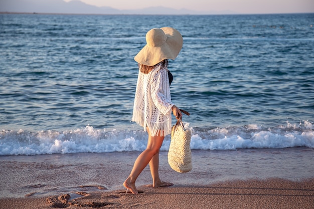 Una niña con un gran sombrero y una bolsa de mimbre camina por la costa del mar. Concepto de vacaciones de verano.