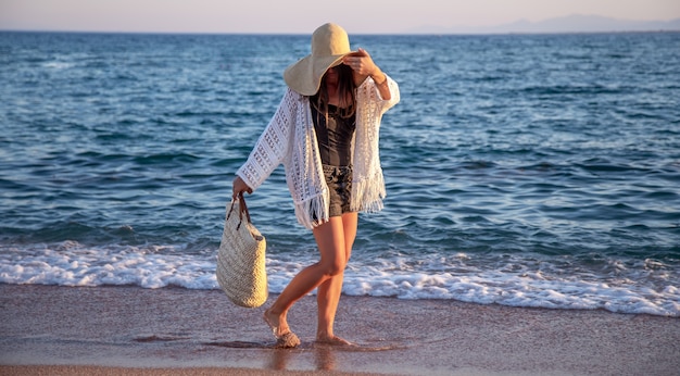 Una niña con un gran sombrero y una bolsa de mimbre camina por la orilla del mar. Concepto de vacaciones de verano.