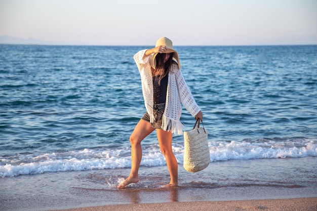 Una niña con un gran sombrero y una bolsa de mimbre camina por la orilla del mar. Concepto de vacaciones de verano.