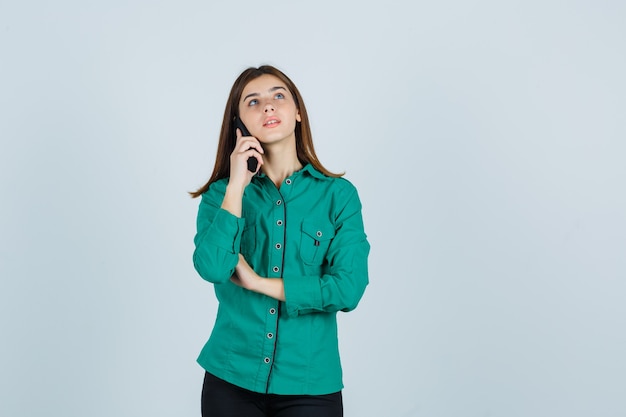 Niña hablando por teléfono, mirando hacia arriba en blusa verde, pantalón negro y mirando enfocado, vista frontal.