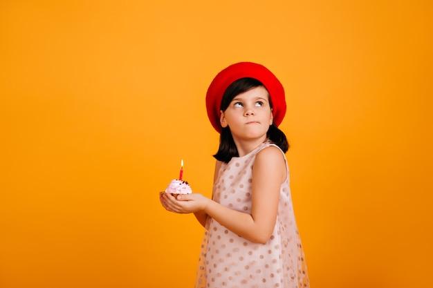 Niña haciendo deseo de cumpleaños. niño morena sosteniendo pastel con vela en la pared amarilla.