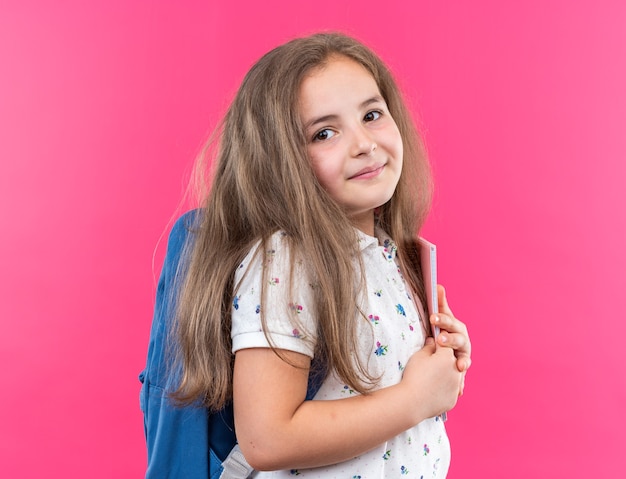 Foto gratuita niña hermosa con el pelo largo con mochila sosteniendo el cuaderno mirando al frente con una sonrisa en la cara feliz de pie sobre la pared rosa