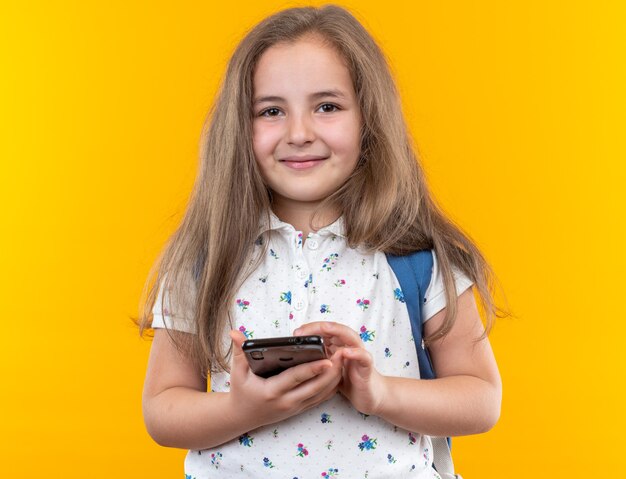 Niña hermosa con el pelo largo con mochila sosteniendo smartphone feliz y alegre sonriente de pie en naranja