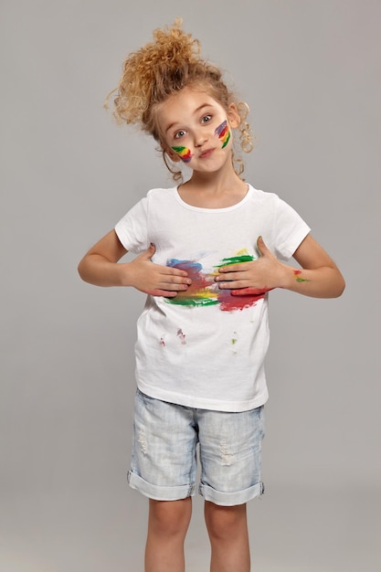 Una niña hermosa que tiene un cabello rubio rizado elegante, que usa una camiseta blanca y pantalones cortos de jeans azules, con los dedos pintados, se está limpiando los brazos en la camiseta, en un fondo gris.