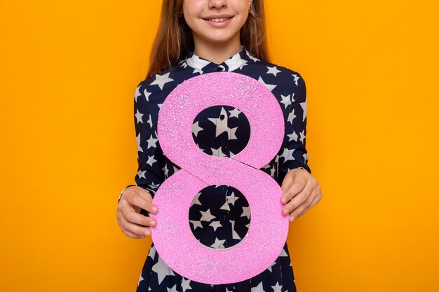 Niña hermosa sonriente en el día de la mujer feliz sosteniendo el número ocho en la cámara aislada en la pared naranja