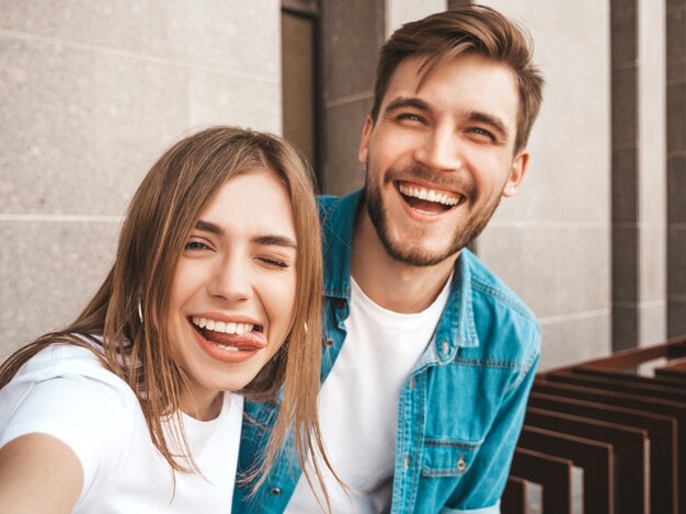 Niña hermosa sonriente y su novio guapo en ropa casual de verano. Familia feliz tomando selfie autorretrato de sí mismos en la cámara del teléfono inteligente. Divirtiéndose en la calle. Muestra lengua