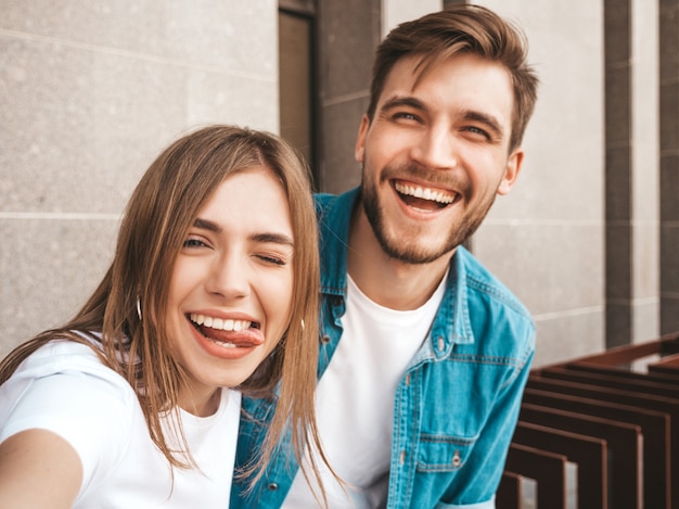 Niña hermosa sonriente y su novio guapo en ropa casual de verano. Familia feliz tomando selfie autorretrato de sí mismos en la cámara del teléfono inteligente. Divirtiéndose en la calle. Muestra lengua