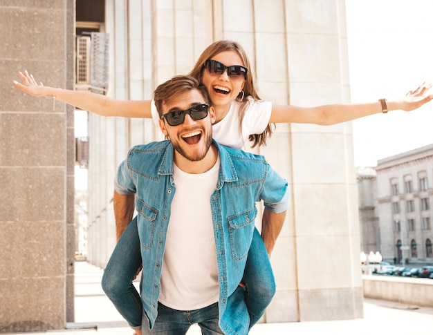 Niña hermosa sonriente y su novio guapo en ropa casual de verano. Hombre con su novia en la espalda y ella levantando las manos.