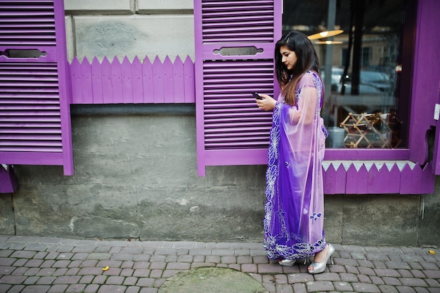 Niña hindú india en sari violeta tradicional posada en la calle contra ventanas moradas con teléfono móvil en las manos