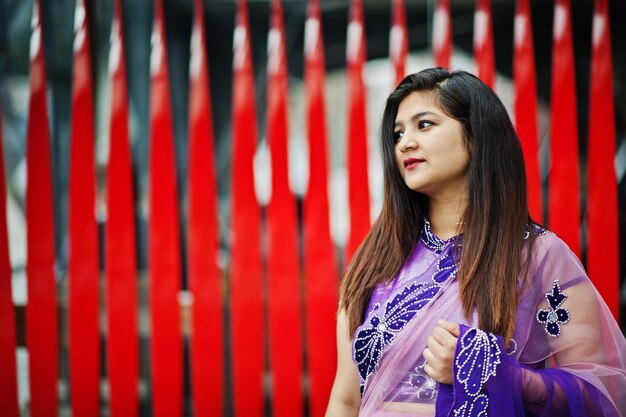 Niña hindú india en el tradicional sari violeta posado en la calle contra el fondo rojo.