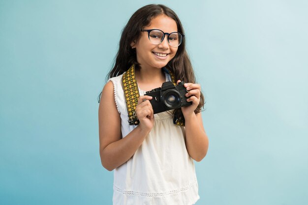 Niña hispana sonriente con cabello castaño largo sosteniendo una cámara digital contra un fondo turquesa