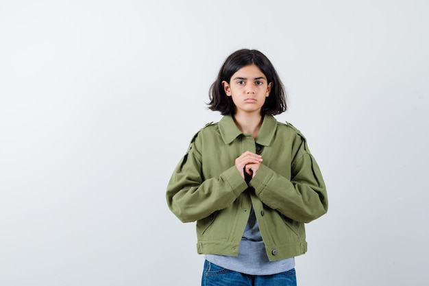 Niña juntando las manos en el pecho en abrigo, camiseta, jeans y luciendo esperanzada. vista frontal.