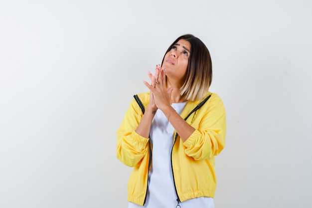 Niña juntando las manos en posición de oración en camiseta blanca, chaqueta amarilla y mirando enfocado, vista frontal.