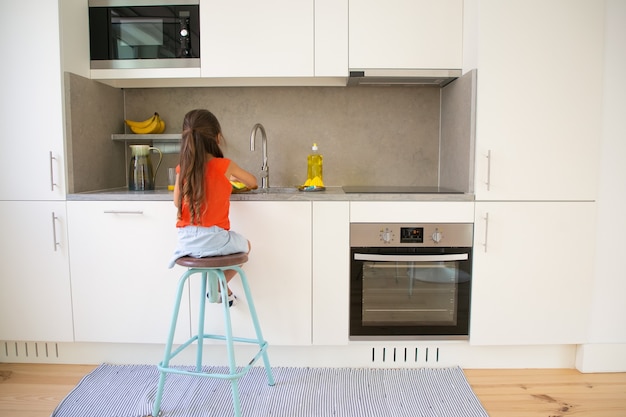 Niña lavando un plato en la cocina por sí misma. Niño sentado en un taburete cerca del fregadero de la cocina, haciendo el trabajo doméstico.