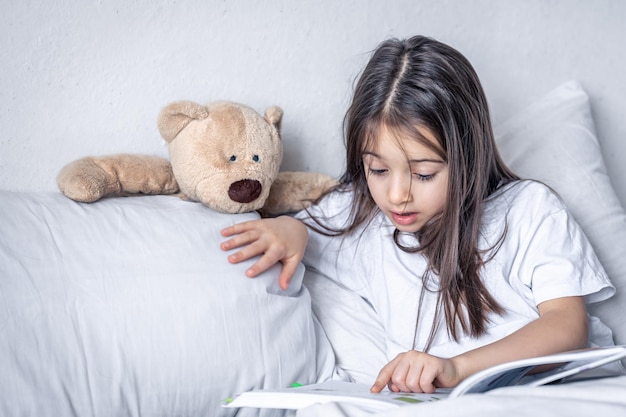 Niña lee un libro con un oso de peluche en la cama por la mañana