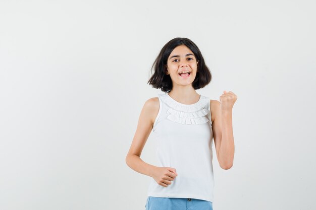 Niña levantando el puño cerrado en blusa blanca, pantalones cortos y mirando feliz. vista frontal.