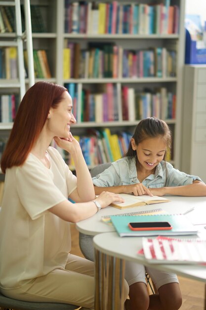 Niña leyendo un libro y viendo a una mujer sentada en la mesa