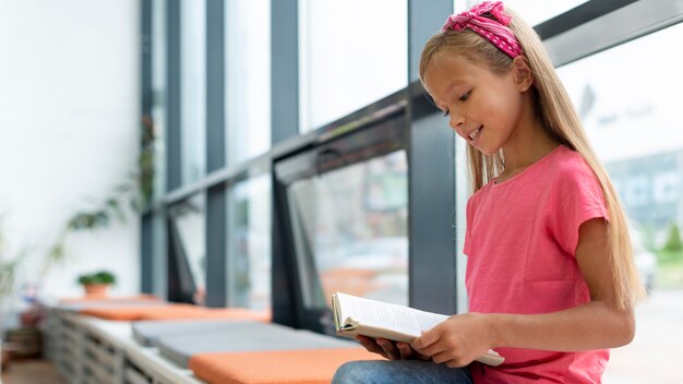 Niña leyendo sentado junto a la ventana con espacio de copia