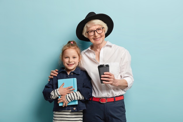 Niña linda con bloc de notas, pasa tiempo libre junto con la abuela, estar de buen humor, beber café para llevar, abrazar. El profesor anciano sonriente abraza a un alumno pequeño, estudia juntos en interiores