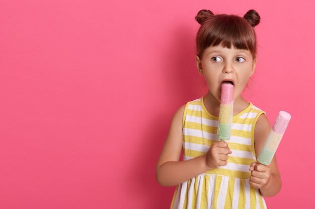 Niña linda con la boca ampliamente abierta mordiendo helado de agua, mirando a un lado mientras posa aislado sobre fondo rosa, copia espacio para publicidad.