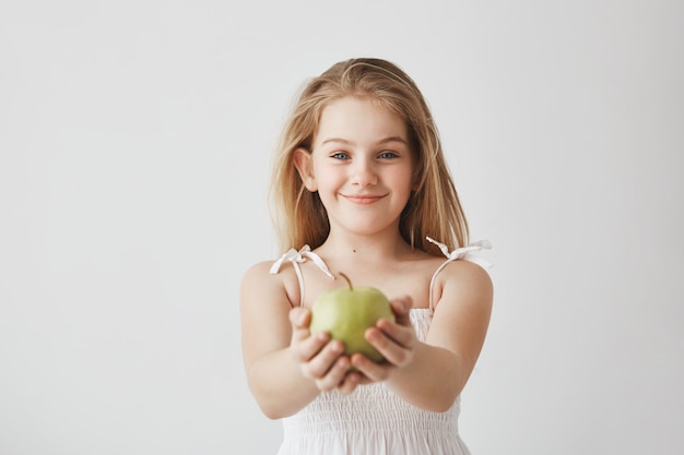 Niña linda con cabello largo rubio y ojos azules en vestido blanco sonriendo, sosteniendo la manzana en las manos y mostrando