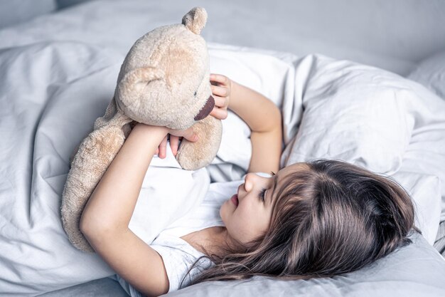 Niña linda en la cama con un oso de peluche temprano en la mañana