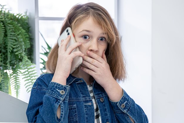 Niña linda en una camisa de mezclilla hablando por teléfono con una cara de sorpresa