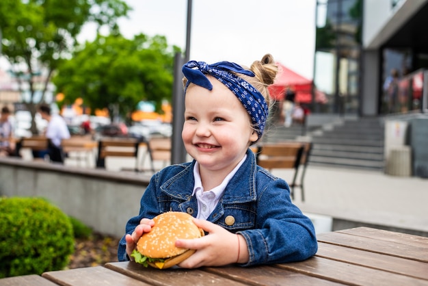 Niña linda comiendo una hamburguesa en un café. Concepto de comida rápida para niños