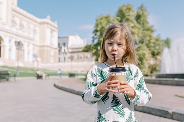 Niña linda concentrada en vestido de verano bebiendo cacao afuera en la luz del sol