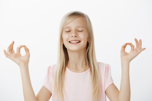 Niña linda meditando y sintiéndose tranquila. retrato de feliz adorable hija amable con cabello rubio, cerrando los ojos con expresión soñadora, levantando las manos con gesto zen, haciendo yoga sobre una pared gris