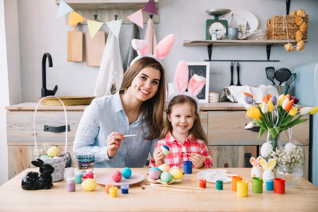 Niña linda en orejas de conejo pintando huevos para Pascua con madre